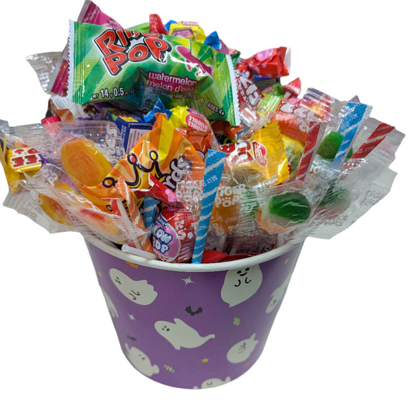 Colorful candy assortment in a purple bucket halloween bucket decorated with cartoon ghosts, on a white surface.