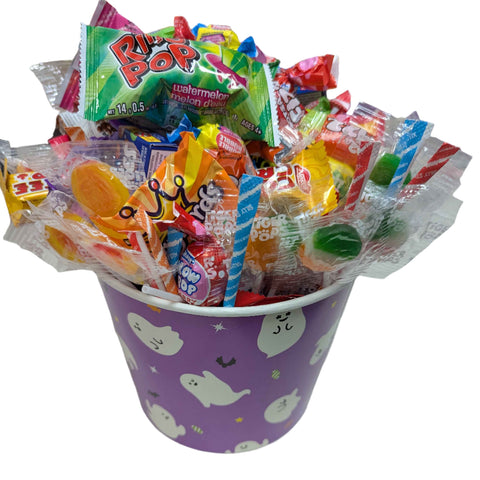 Colorful candy assortment in a purple bucket halloween bucket decorated with cartoon ghosts, on a white surface.