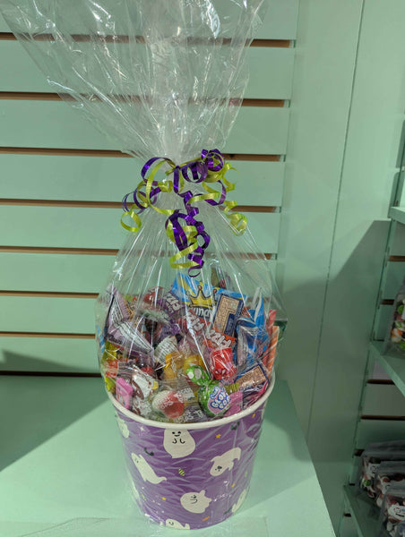Purple halloween bucket decorated with cartoon ghosts, filled with colorful candies and wrapped in clear cellophane with ribbons against a light green wall.
